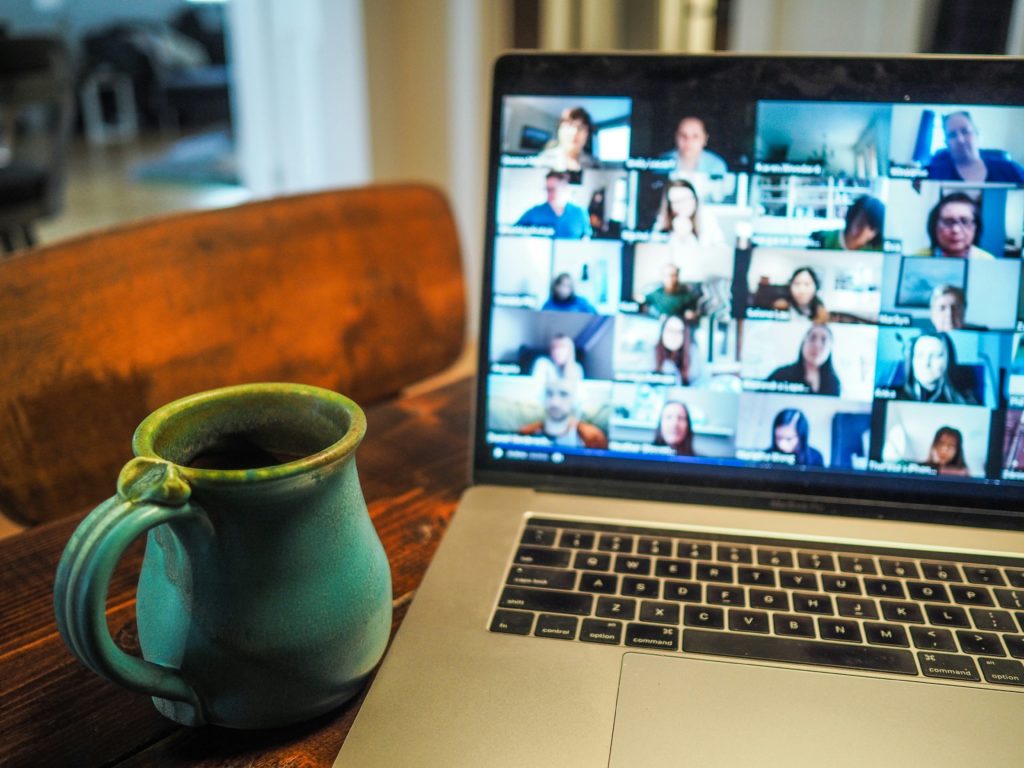 Laptop showing a video call with multiple participants on a virtual meeting platform, sitting on a wooden table next to a green mug, representing a remote work setup.