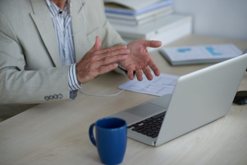 Man in a beige suit gesturing while on a video call with a laptop. Documents and a blue mug are on the desk, conveying a professional tone.